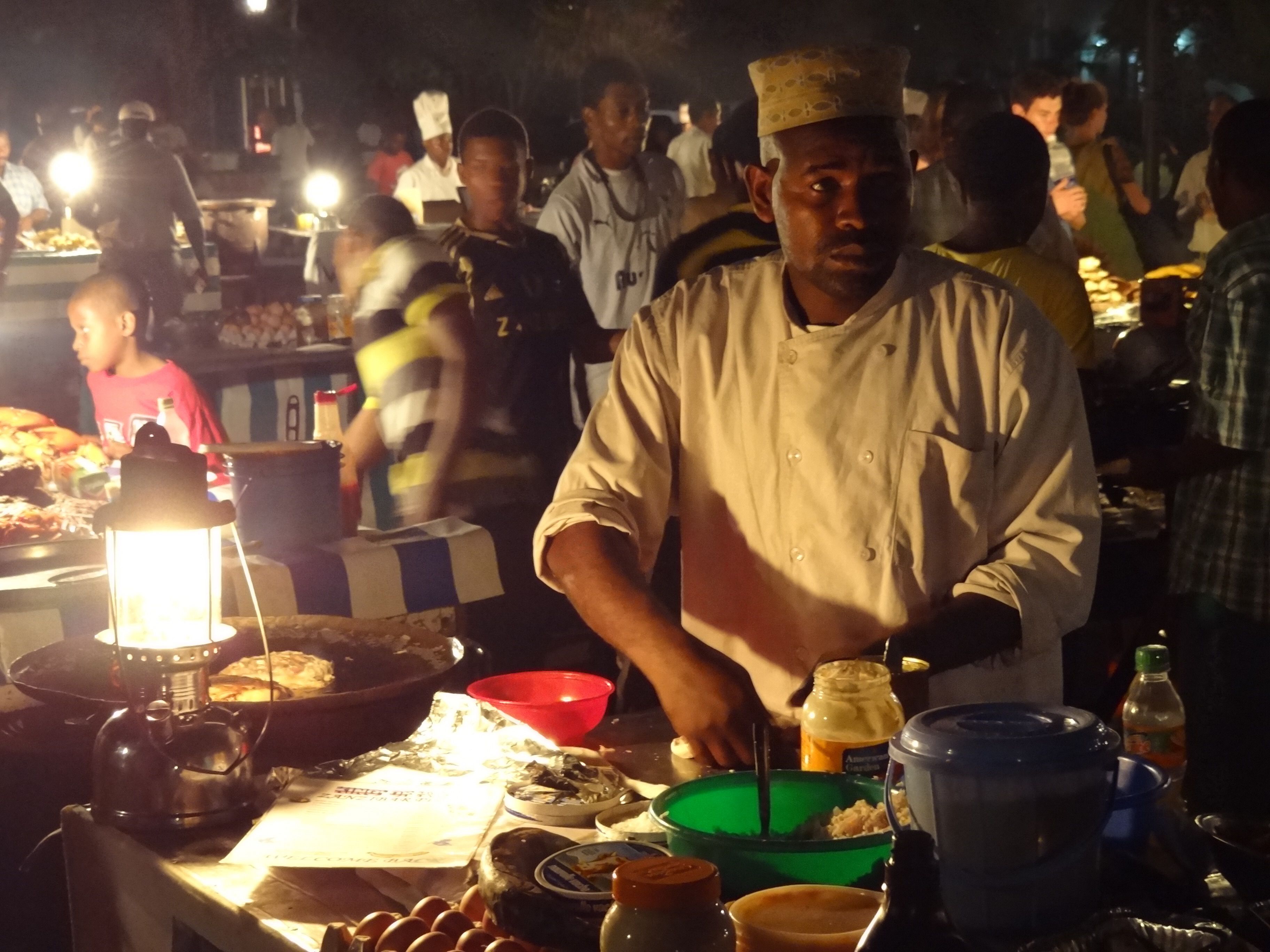 A cook at a lantern-lit food stall at the Forodhani Gardens night market on the Stone Town seafront, a hurricane lamp glowing over a charcoal pan of food in the foreground and a crowd of diners and vendors behind him in the dark.