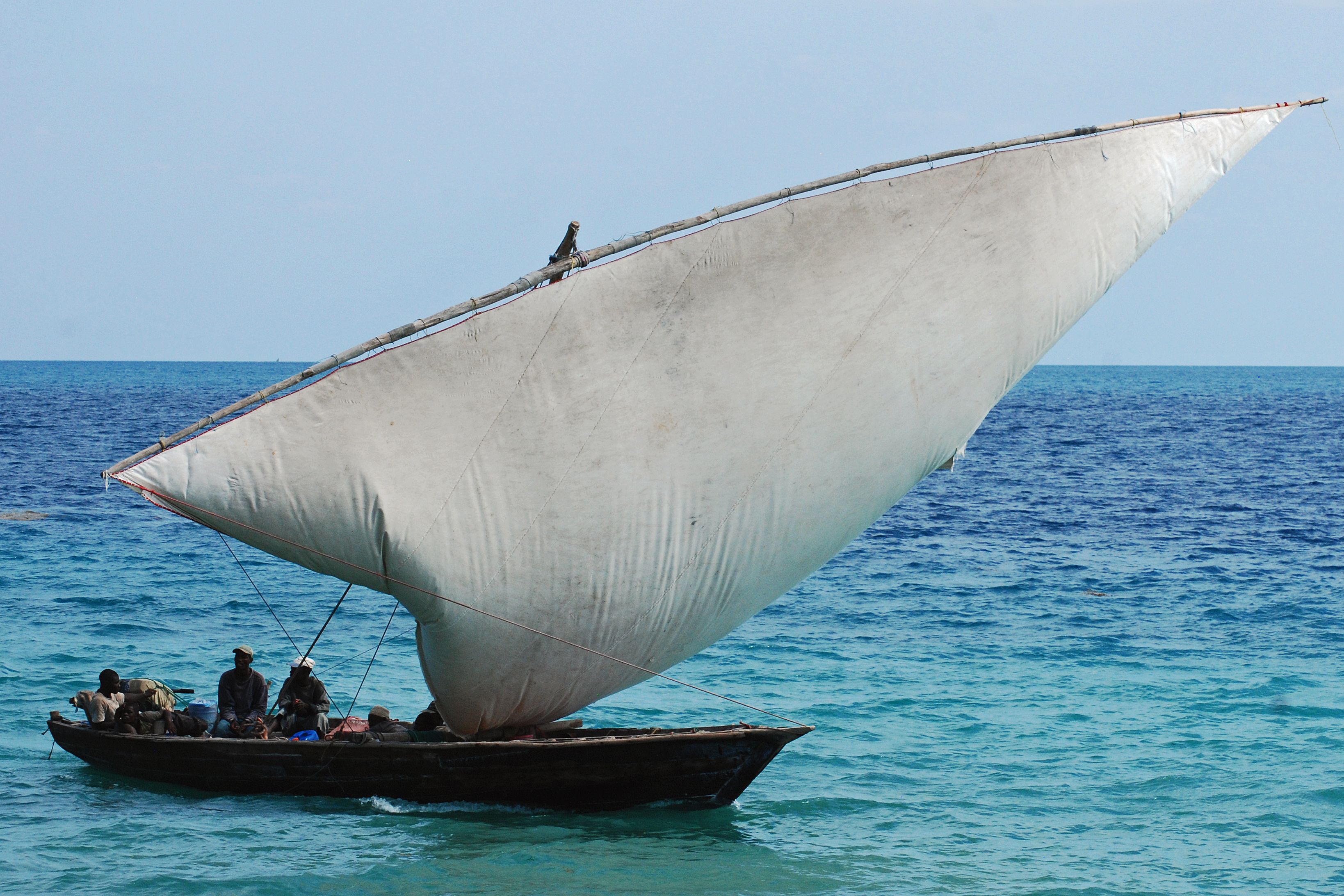 A traditional wooden Zanzibar dhow under a full white lateen sail, carrying several men across turquoise-to-deep-blue water under a clear bright sky.