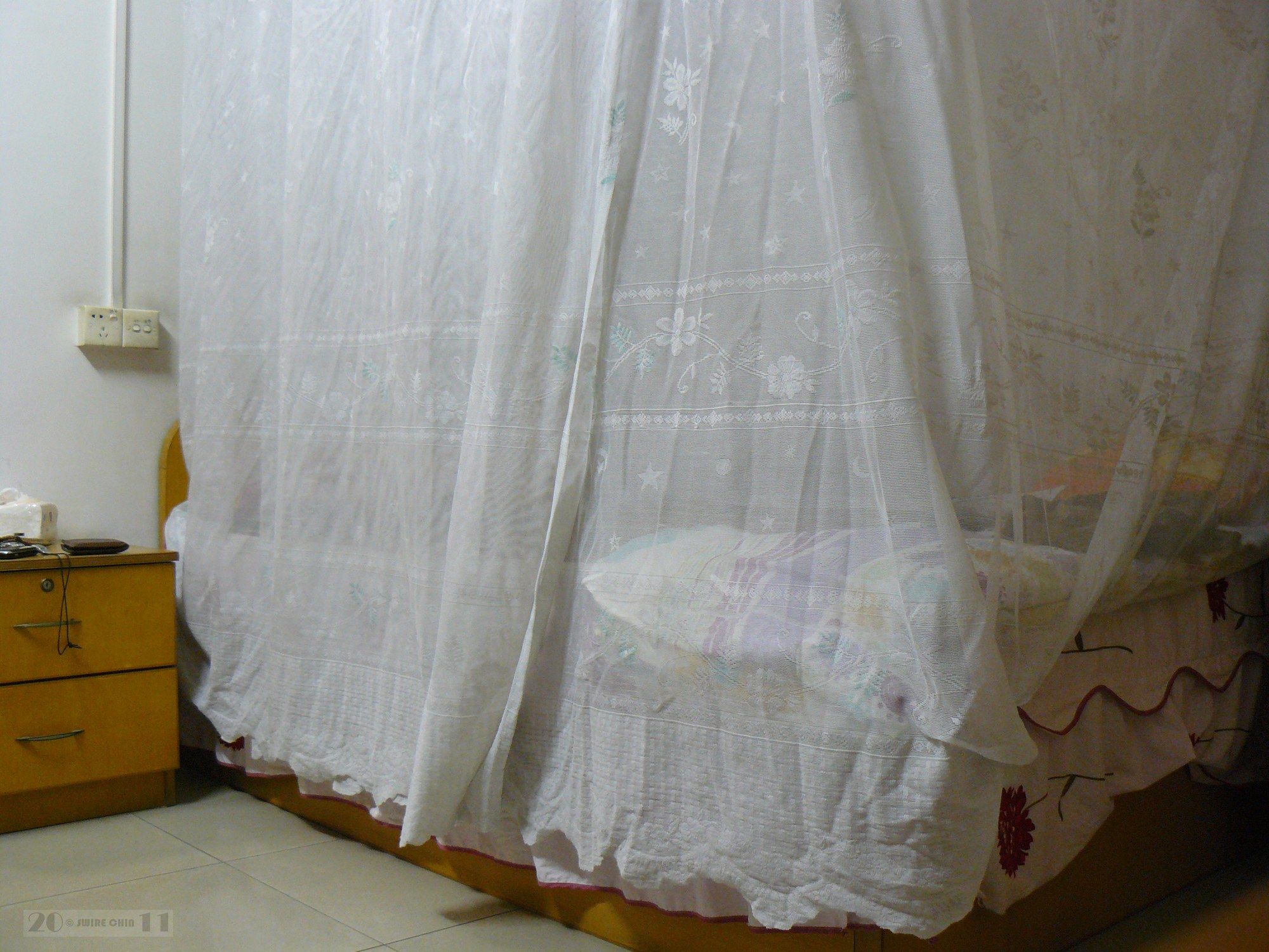 A white mosquito net draped over a bed next to a wooden bedside table in a sunlit room