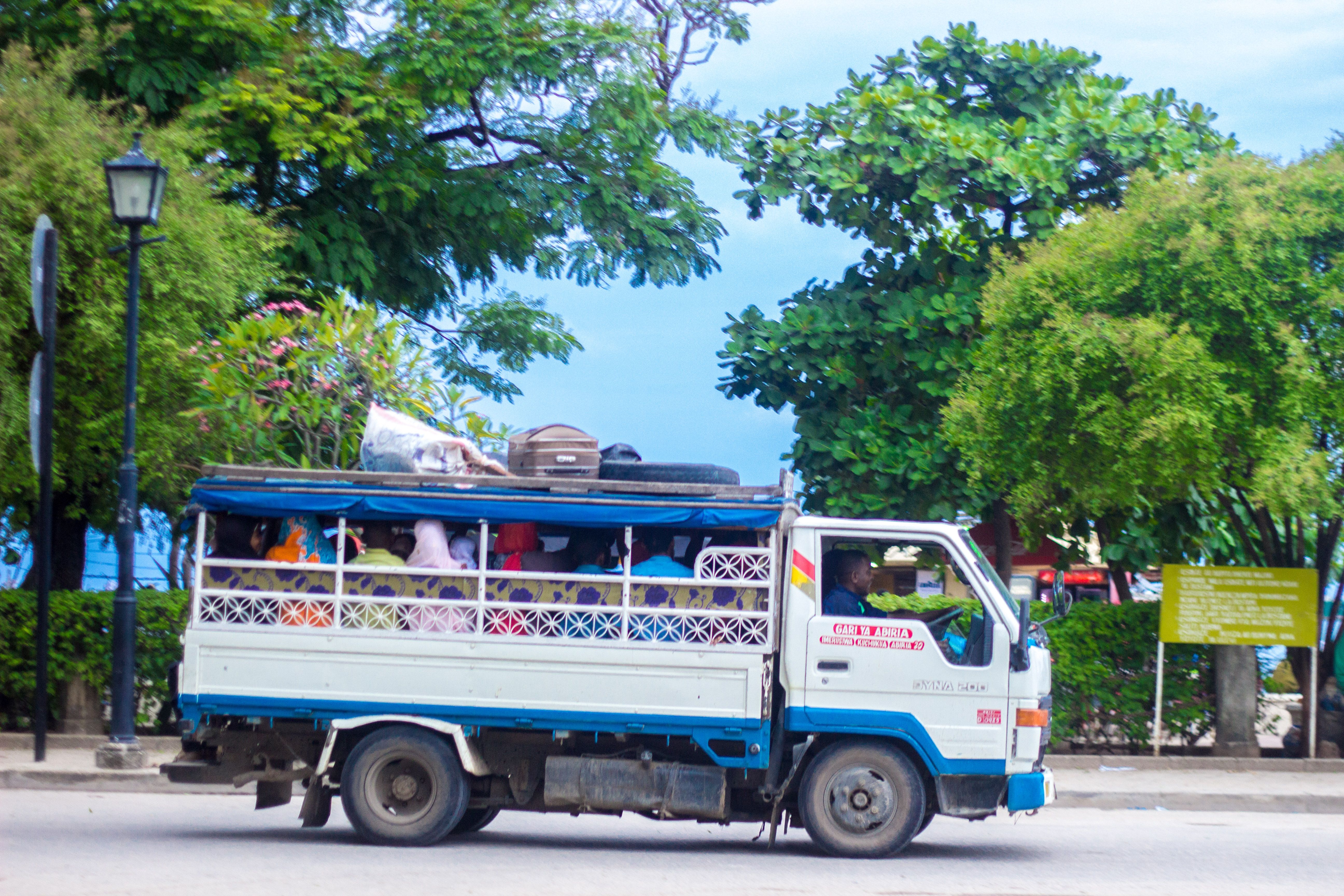 A converted Toyota Dyna truck dala-dala with brightly patterned side panels and luggage stacked on the roof rack, packed with seated passengers, driving down a tree-lined Stone Town street.