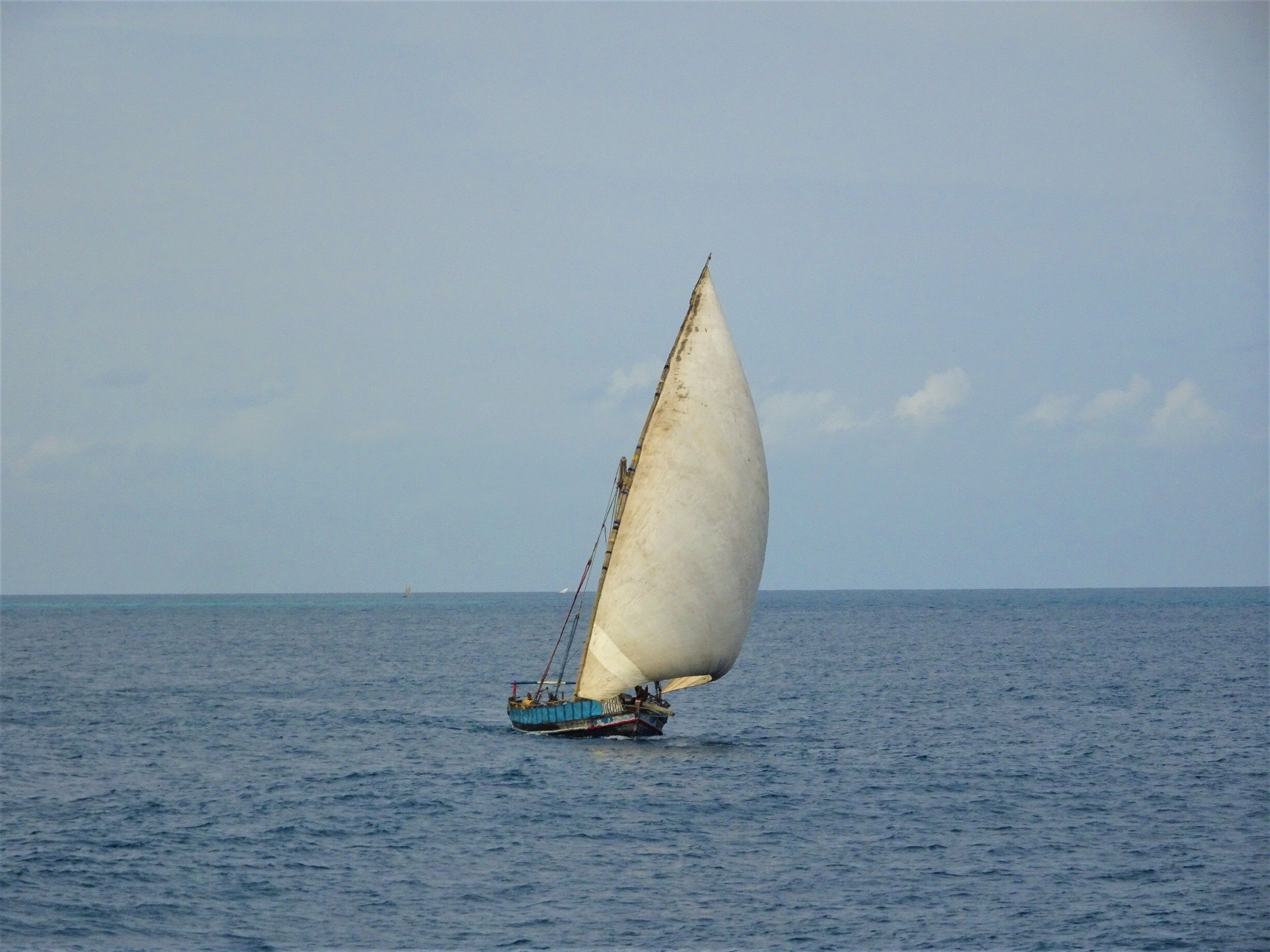 A traditional wooden dhow under full sail on the deep-blue Indian Ocean off Zanzibar, its white lateen sail full of wind and crew aboard the blue-painted hull