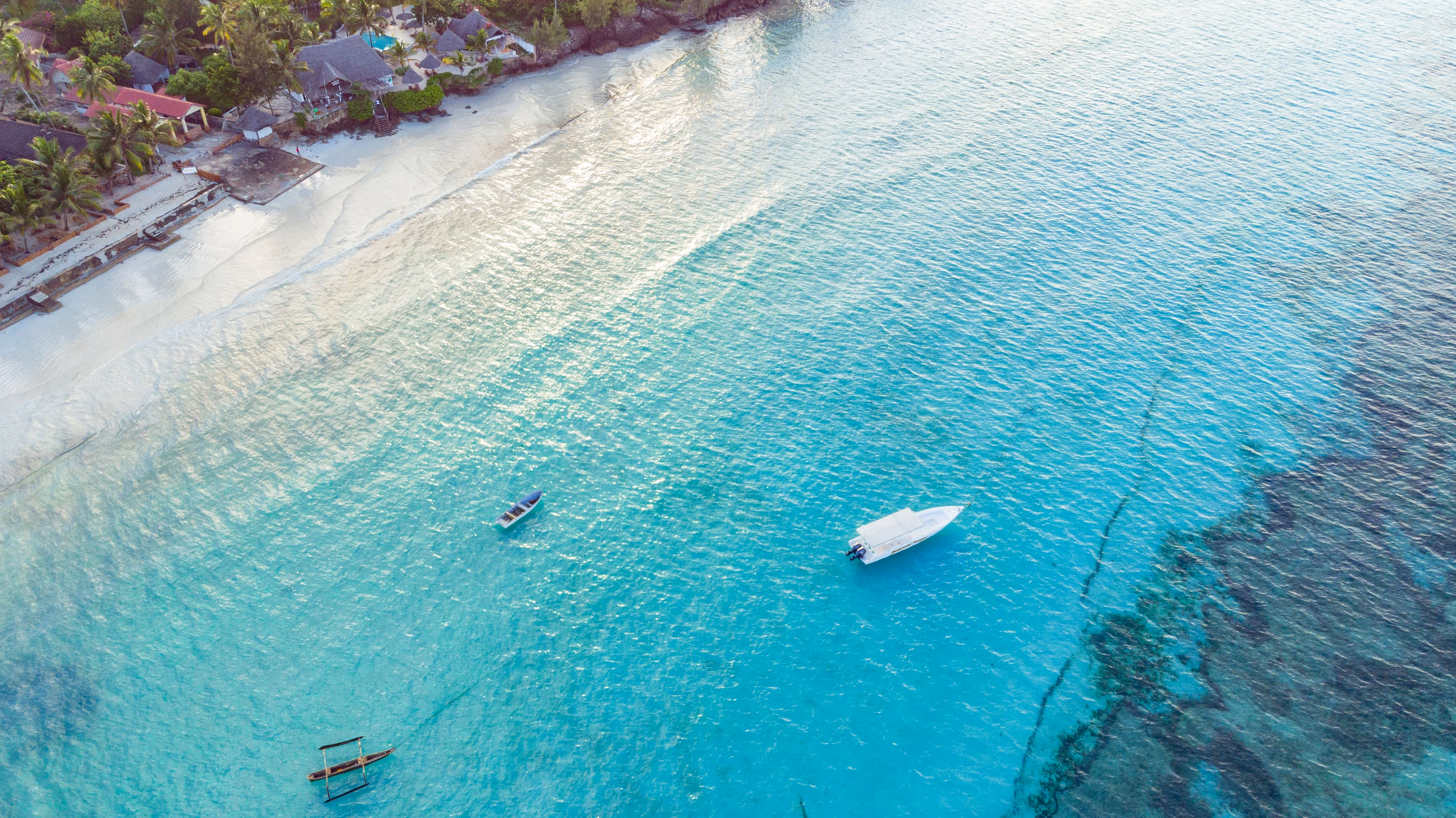 An aerial view looking down over a white-sand Zanzibar beach lined with palm trees and thatched-roof bungalows, where clear turquoise shallows stretch out to deeper blue water and a traditional wooden outrigger boat and two small boats rest on the calm sea.