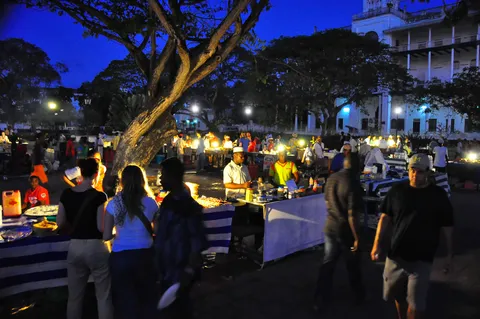 Forodhani Gardens night food market at dusk in Stone Town, Zanzibar: lantern-lit vendor stalls with white tablecloths and a charcoal grill, evening crowds under a large tree, and the lit seafront buildings behind.