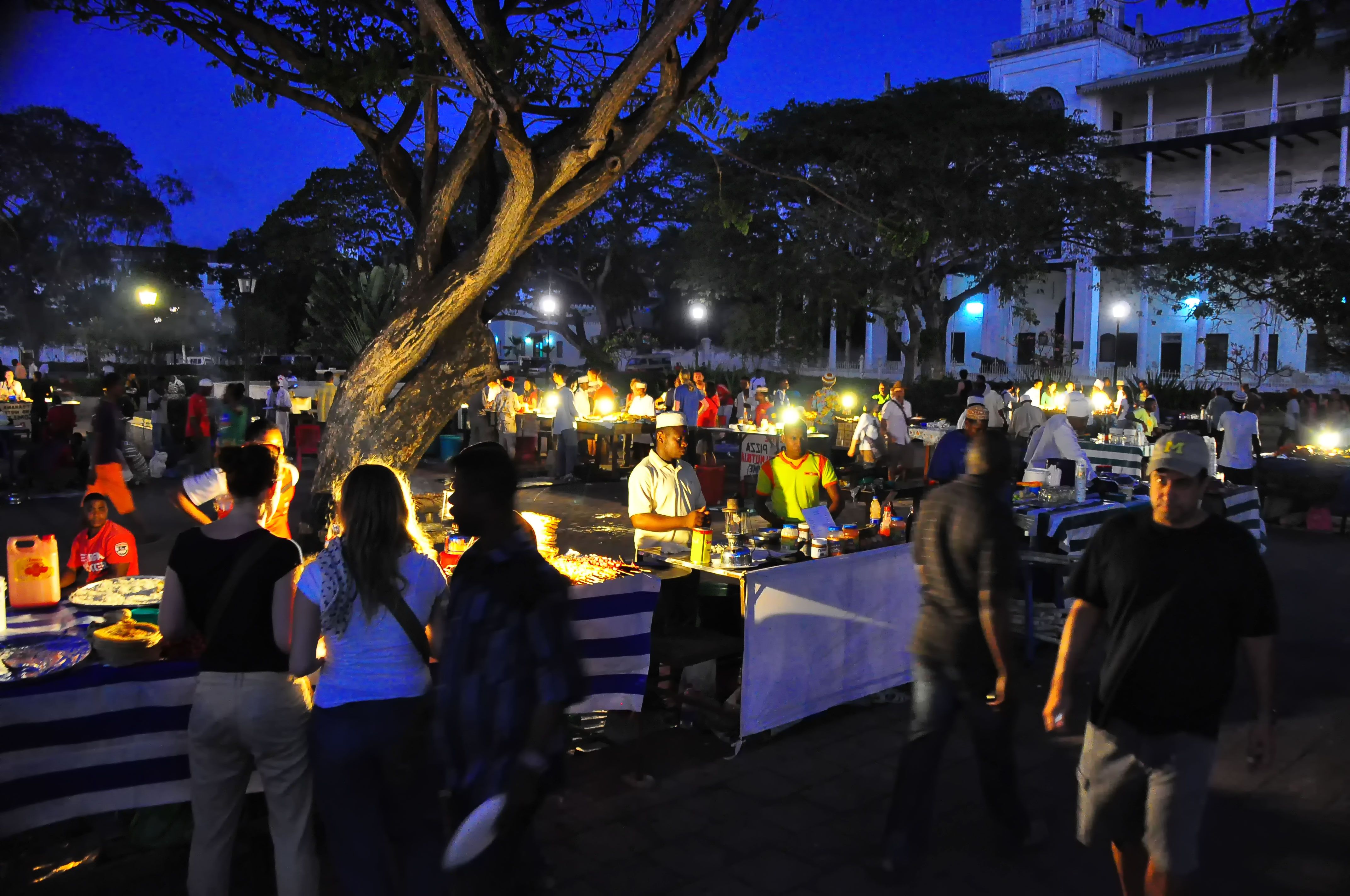 Forodhani Gardens night food market at dusk in Stone Town, Zanzibar: lantern-lit vendor stalls with white tablecloths and a charcoal grill, evening crowds under a large tree, and the lit seafront buildings behind.