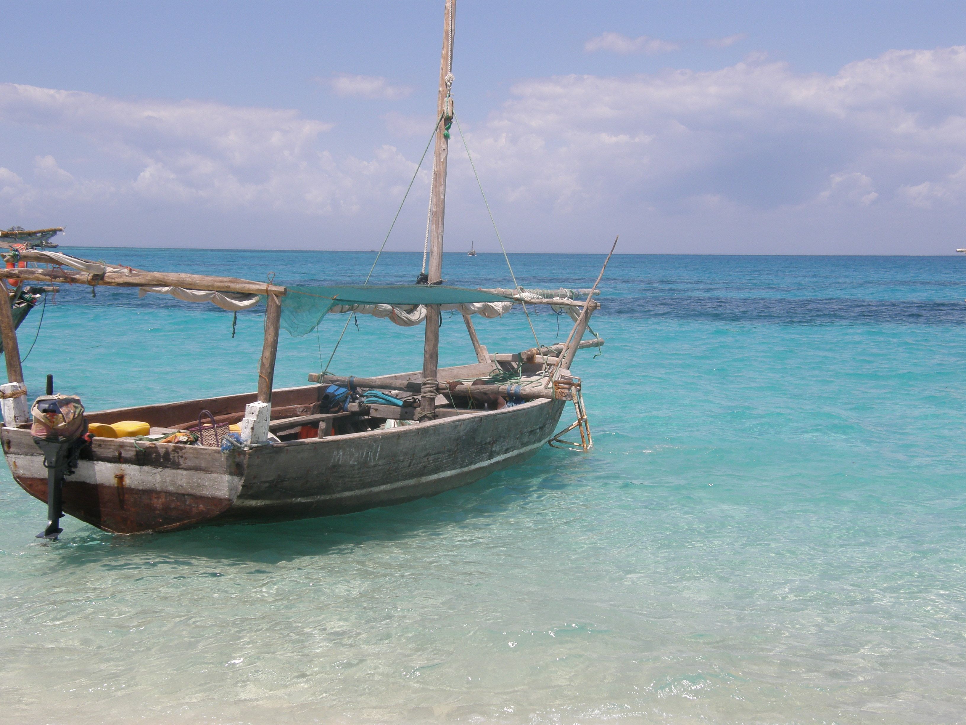 A traditional wooden dhow anchored in clear turquoise shallows off a bright sandy shore near Kwale Island in Menai Bay, Zanzibar, under a bright blue sky
