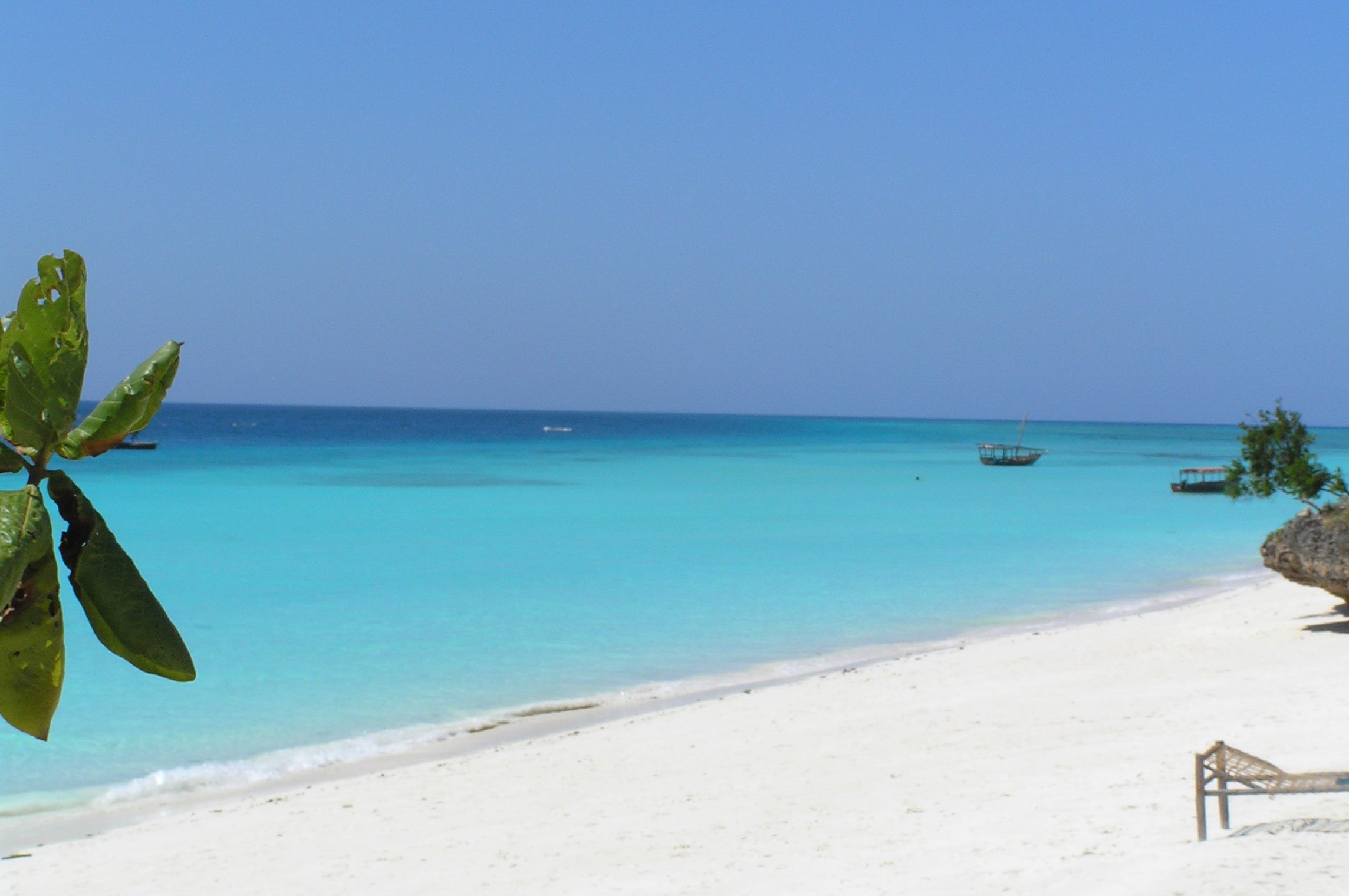 A sunny Nungwi beach on Zanzibar's north coast with white sand, bright turquoise water, and two traditional wooden dhows moored offshore under a clear blue sky