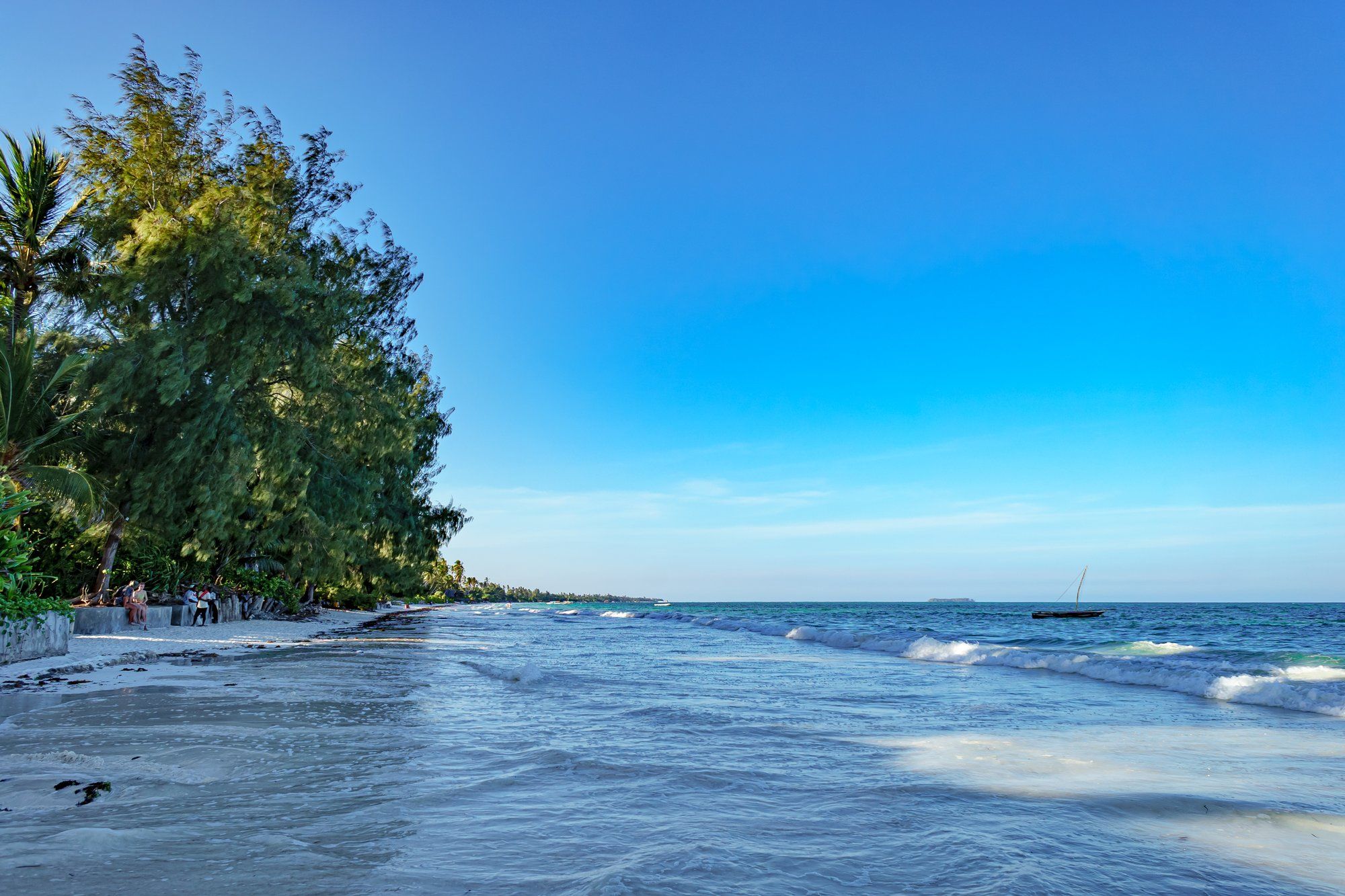 The calm turquoise sea on Zanzibar's north coast at Nungwi in warm late-afternoon light, a wooden dhow anchored near the shore on the right, casuarina and palm trees leaning over the white sand on the left, and a small islet on the horizon.