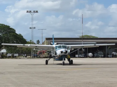 A single-engine Cessna Caravan turboprop parked on the sunlit apron at Zanzibar's airport, with a ground handler, hangars and palms behind under a bright, partly clouded sky.