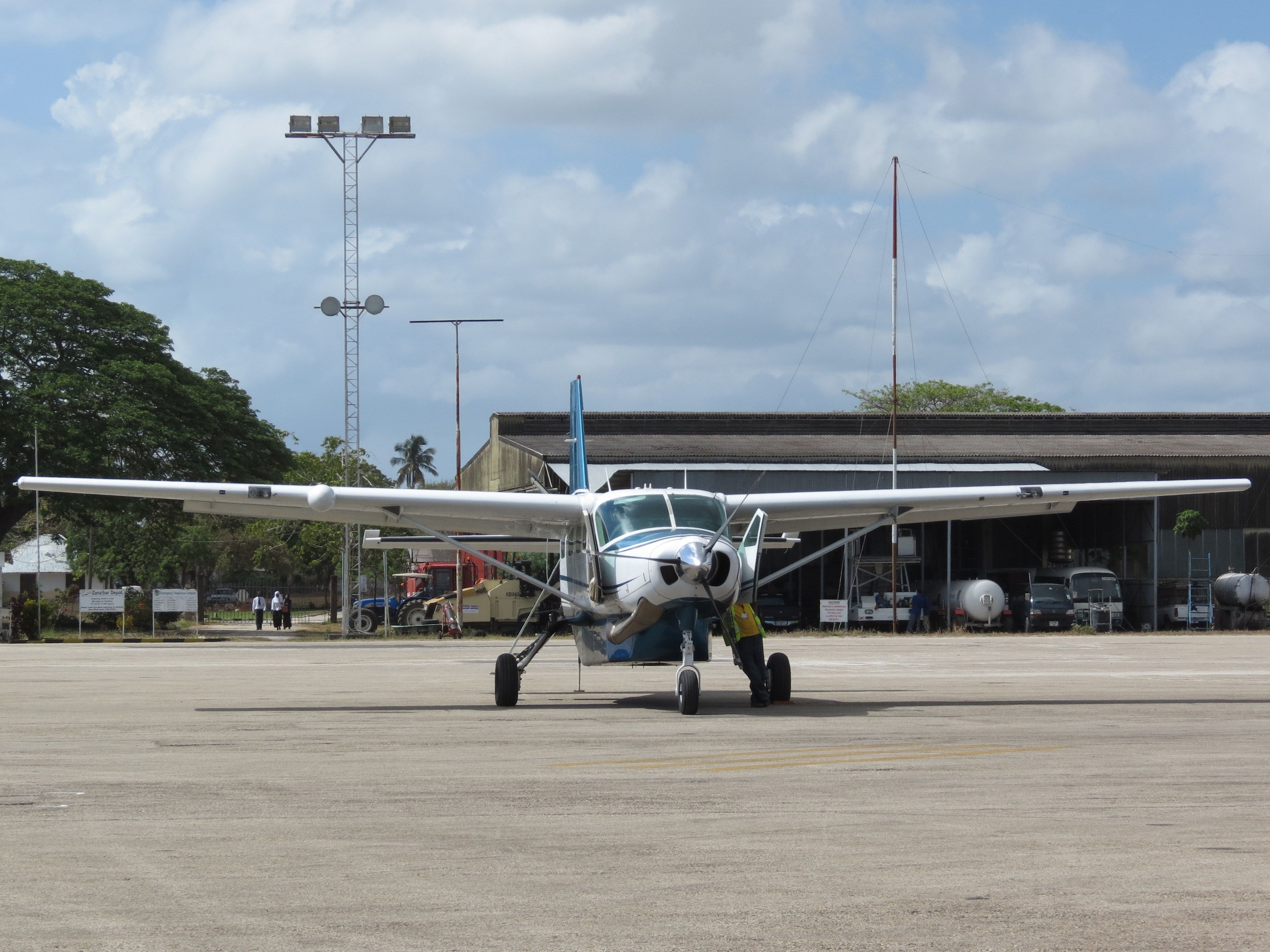 A single-engine Cessna Caravan turboprop parked on the sunlit apron at Zanzibar's airport, with a ground handler, hangars and palms behind under a bright, partly clouded sky.