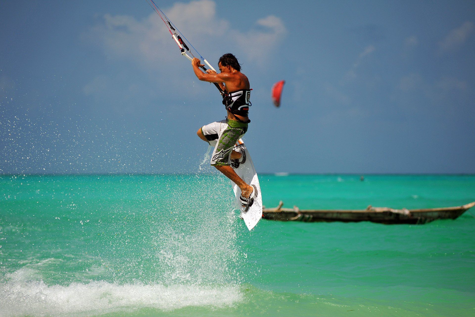 A kitesurfer jumping above a shallow turquoise Zanzibar lagoon, kite overhead and a wooden dhow on the water behind