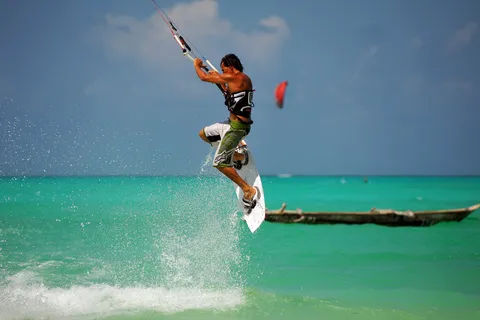 A kitesurfer jumping above a shallow turquoise Zanzibar lagoon, kite overhead and a wooden dhow on the water behind