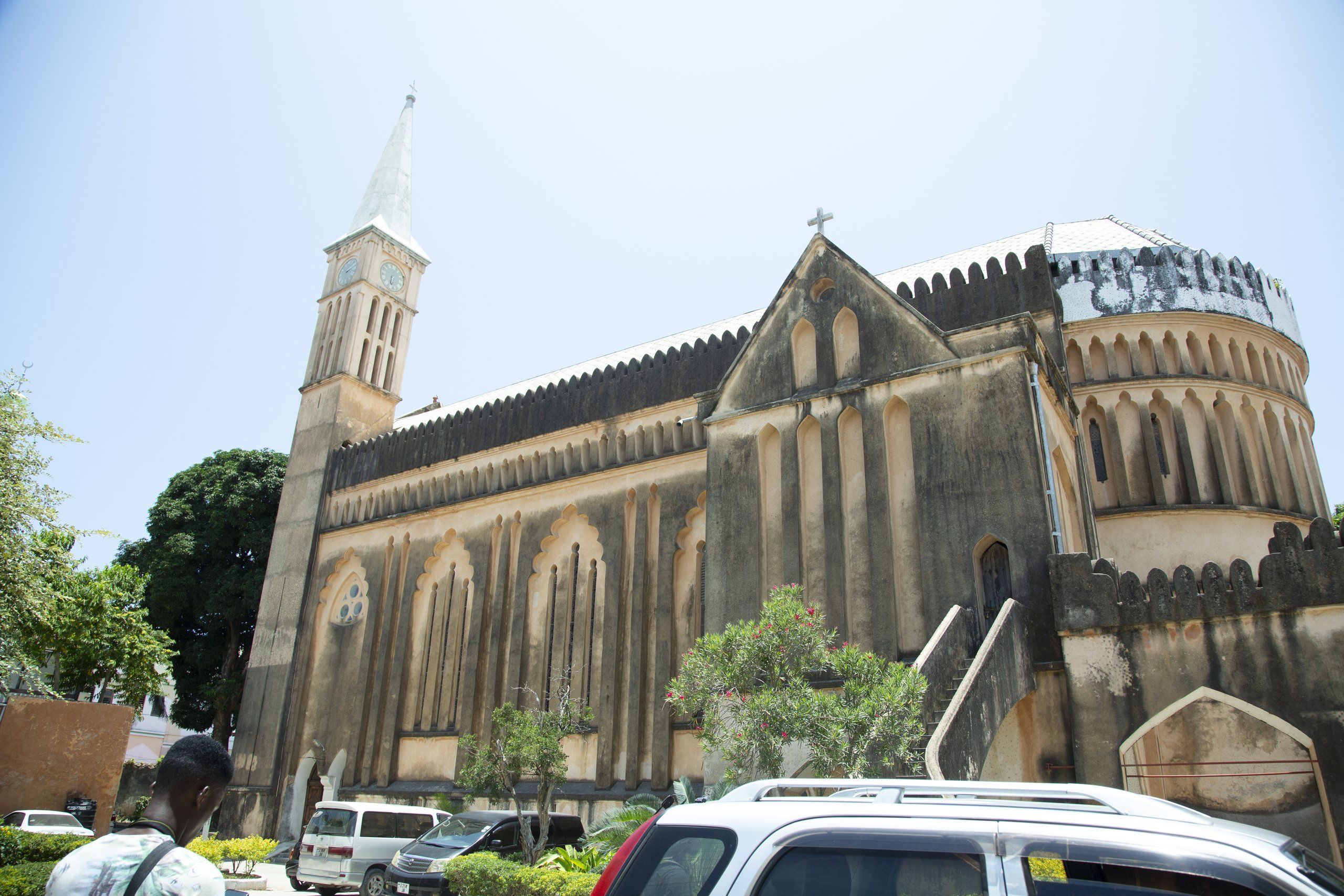 Christ Church Anglican Cathedral in Stone Town, Zanzibar, its whitewashed clock tower and slender spire rising on the left above a weathered grey coral-stone facade with tall pointed-arch buttresses and a cross on the gable, the rounded apse at the right, with a tree, low shrubs and a few parked cars in the foreground under a pale hazy sky.
