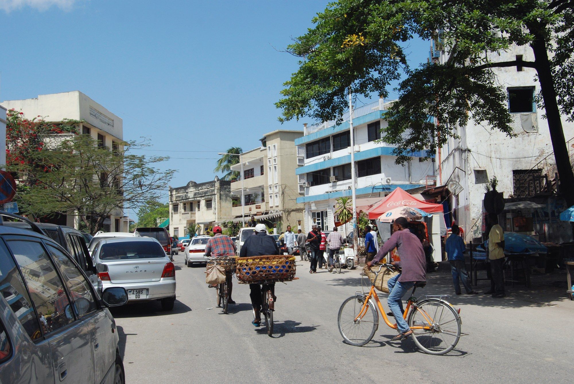 A bright, sunny street in Stone Town, Zanzibar, with people cycling and walking past shops and parked cars beside coral-stone buildings under a blue sky
