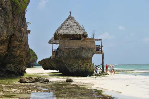 The Rock restaurant, a thatched hut on a single coral outcrop beside a tall cliff at low tide off Pingwe on Zanzibar's Michamvi peninsula, with the exposed reef flat and turquoise sea behind.