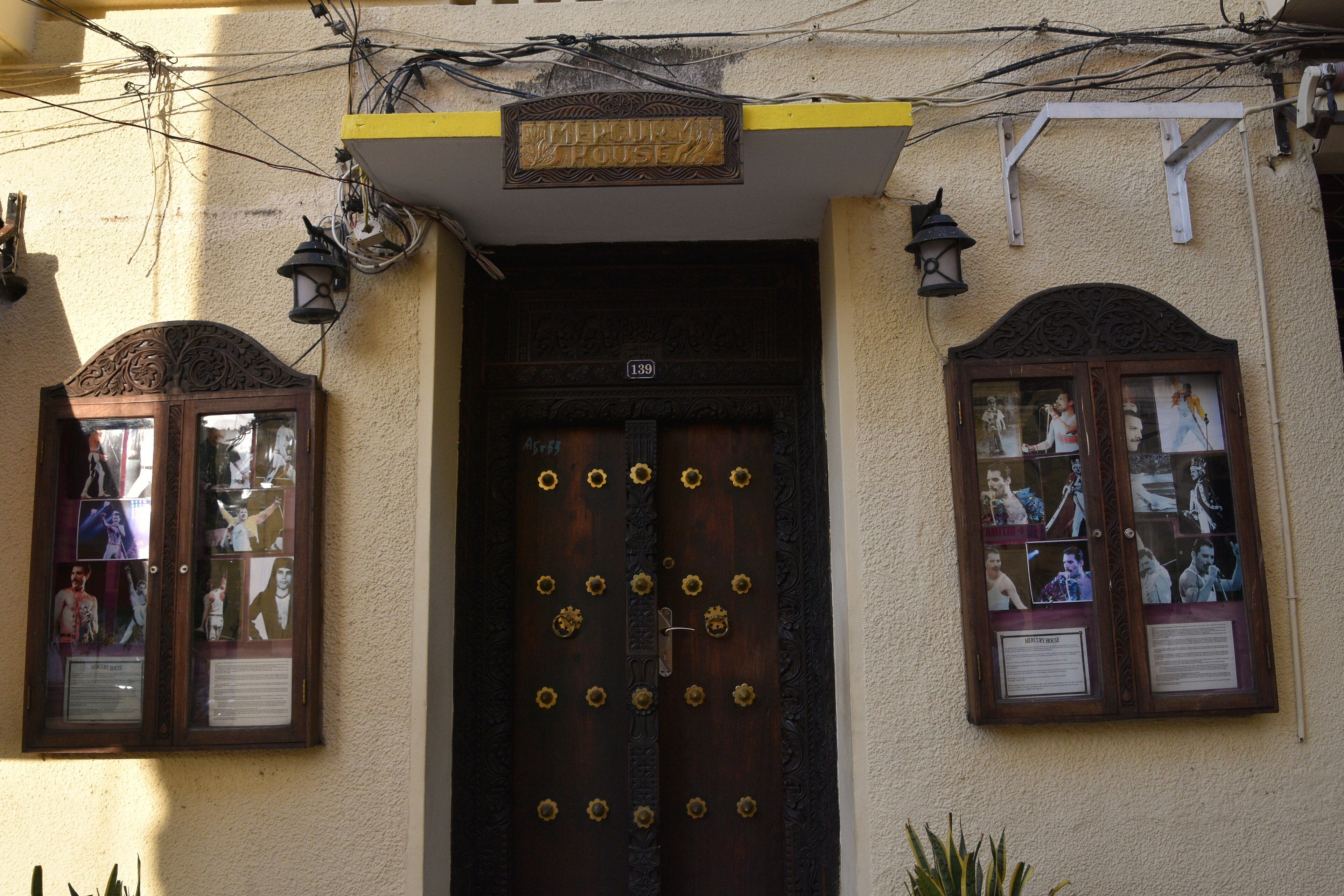 The entrance of Mercury House in Stone Town, the Freddie Mercury Museum: a heavy carved wooden Zanzibar door beneath a brass 'Mercury House' sign on a pale coral-stone facade, flanked by two glazed cases of large photographs of the Queen singer beside the doorway.
