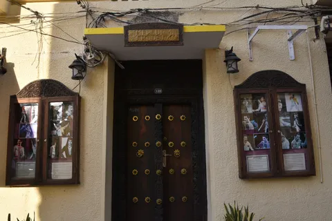 The entrance of Mercury House in Stone Town, the Freddie Mercury Museum: a heavy carved wooden Zanzibar door beneath a brass 'Mercury House' sign on a pale coral-stone facade, flanked by two glazed cases of large photographs of the Queen singer beside the doorway.