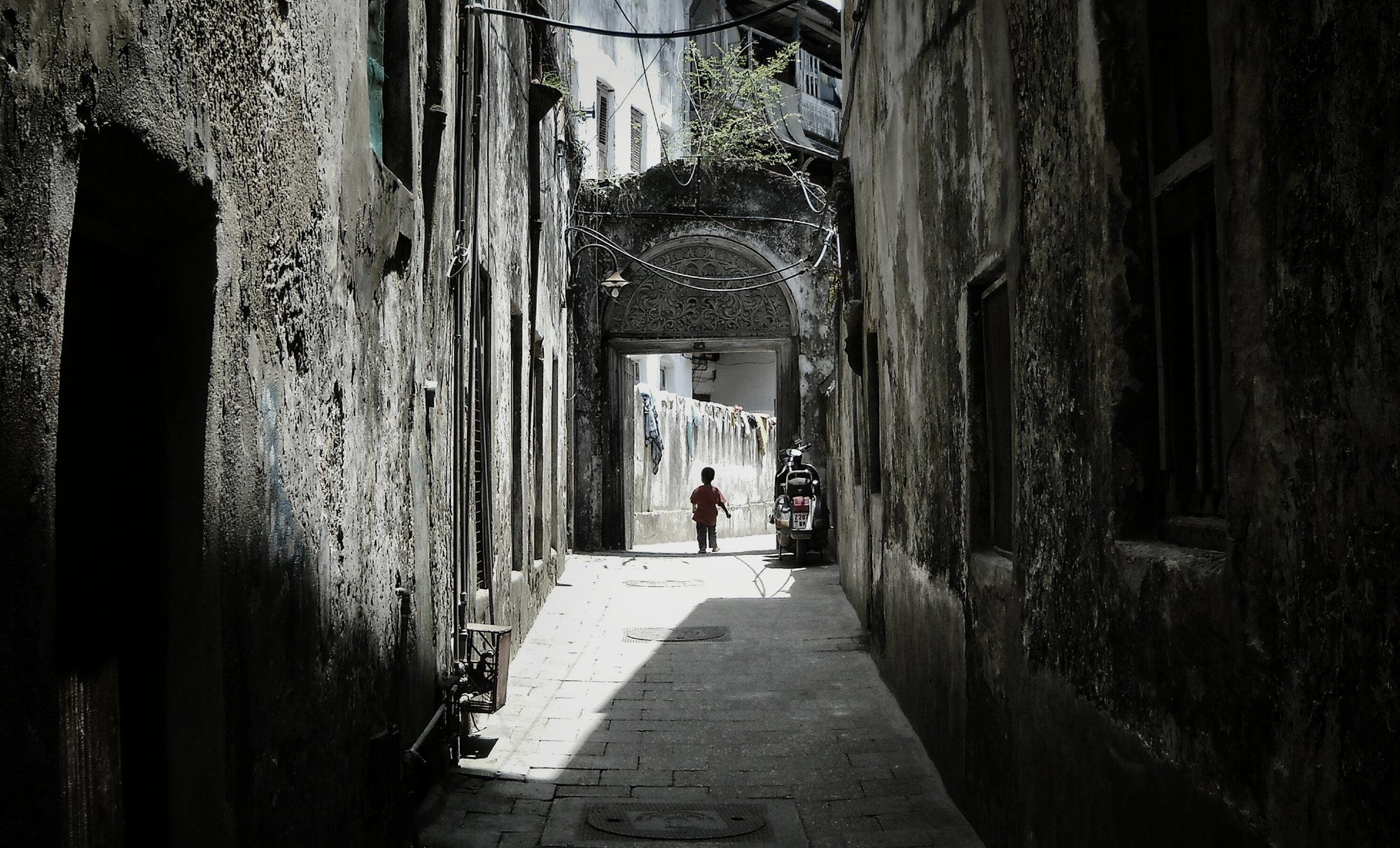 A narrow, shaded coral-stone alley in Stone Town, Zanzibar, leading to a carved-arch doorway with laundry strung overhead, the maze of lanes you are meant to get a little lost in.