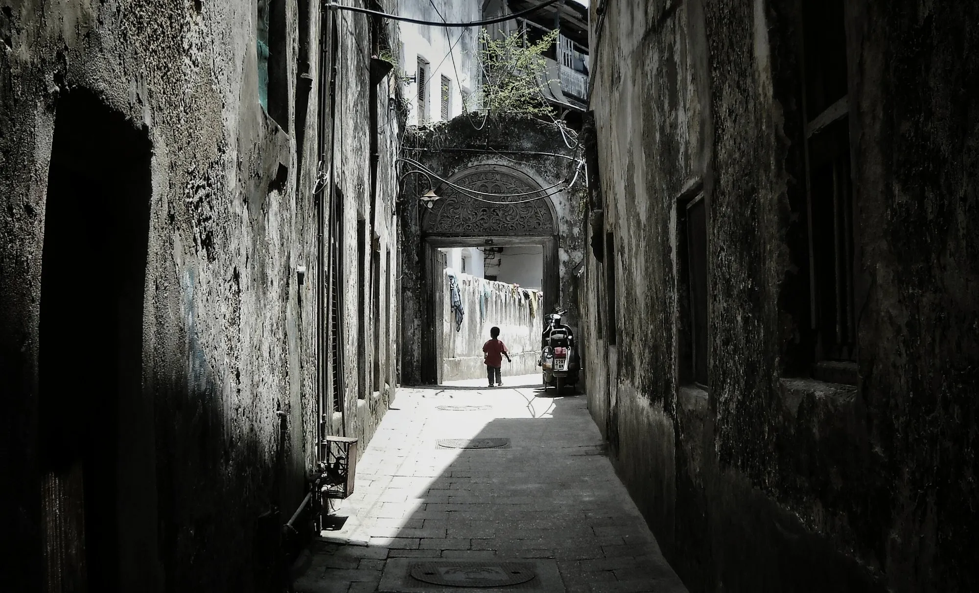 A narrow, shaded coral-stone alley in Stone Town, Zanzibar, leading to a carved-arch doorway with laundry strung overhead, the maze of lanes you are meant to get a little lost in.
