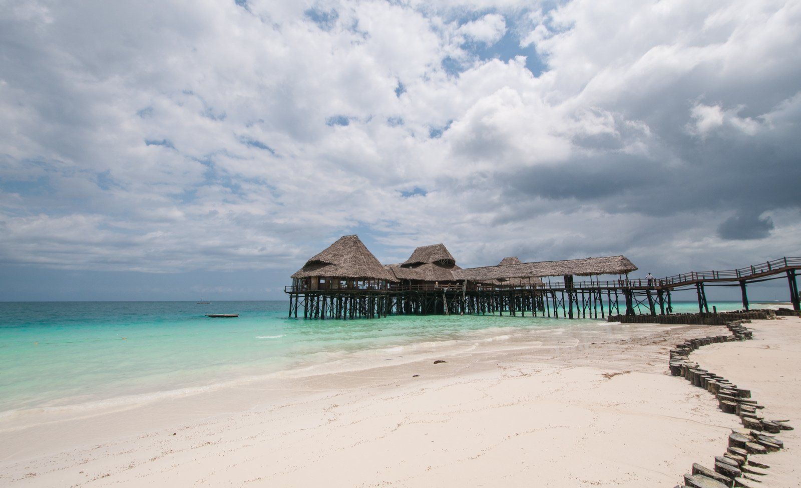A thatched-roof beach bar and jetty on stilts reaching over the clear turquoise water at Kendwa on Zanzibar's north tip, wide white sand in the foreground, where deep water and calm sunsets make the north the easy swim-anytime base.