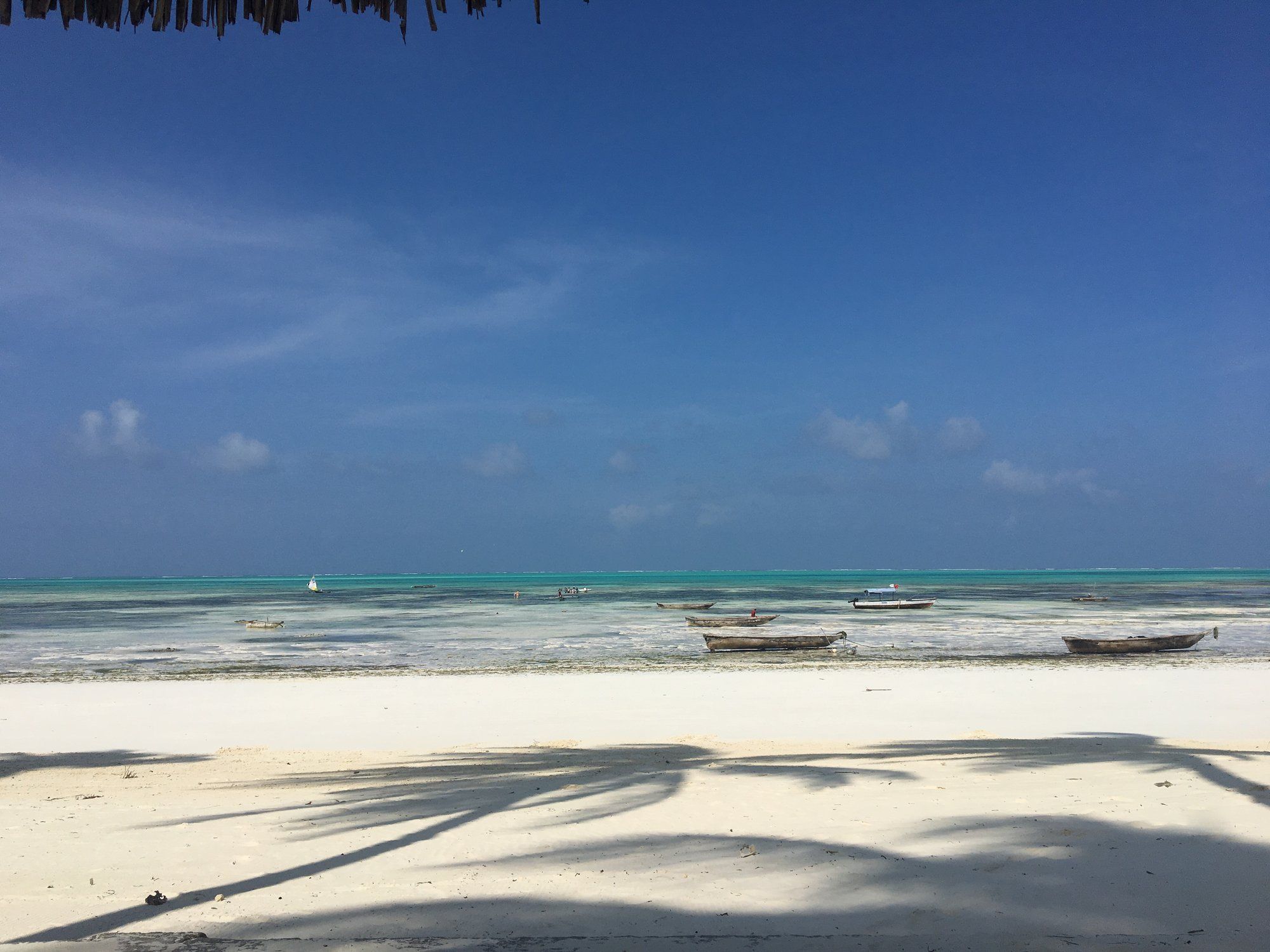 Calm, clear turquoise shallows over pale sand at low tide on Zanzibar's coast, a couple of small wooden boats resting on the flat, the glassy clarity the calm season is known for.