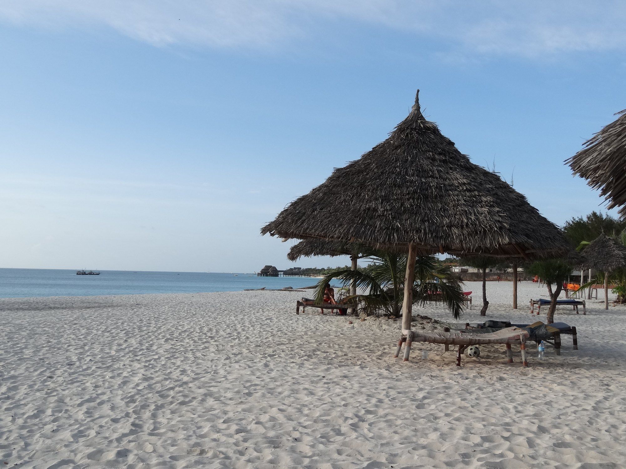 A calm, palm-shaded beach with thatched sun umbrellas and wooden loungers on the pale sand at Kendwa on Zanzibar's north coast, where the sea stays swimmable at any tide, the easy post-safari base.