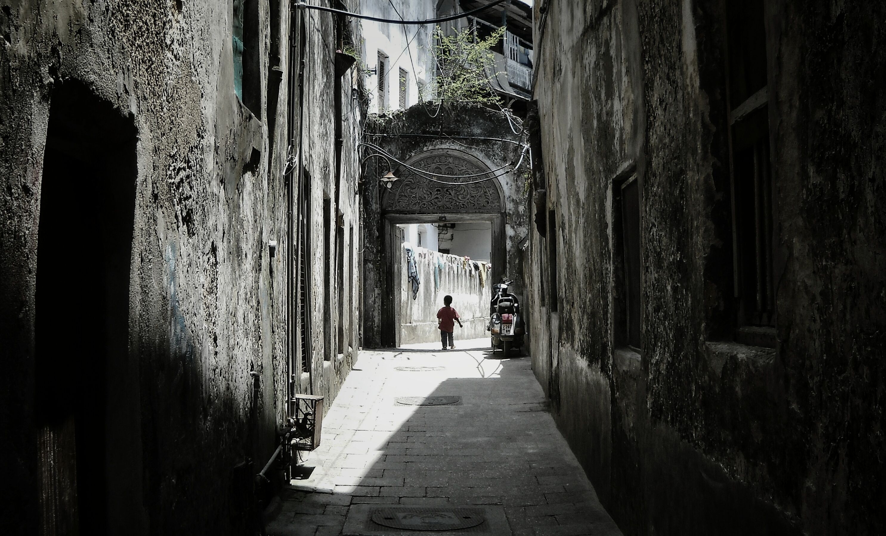 A narrow whitewashed alley in Stone Town, Zanzibar, leading to a carved-arch doorway with laundry strung overhead.