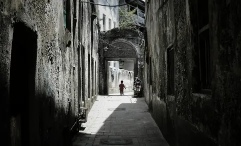 A narrow whitewashed alley in Stone Town, Zanzibar, leading to a carved-arch doorway with laundry strung overhead.