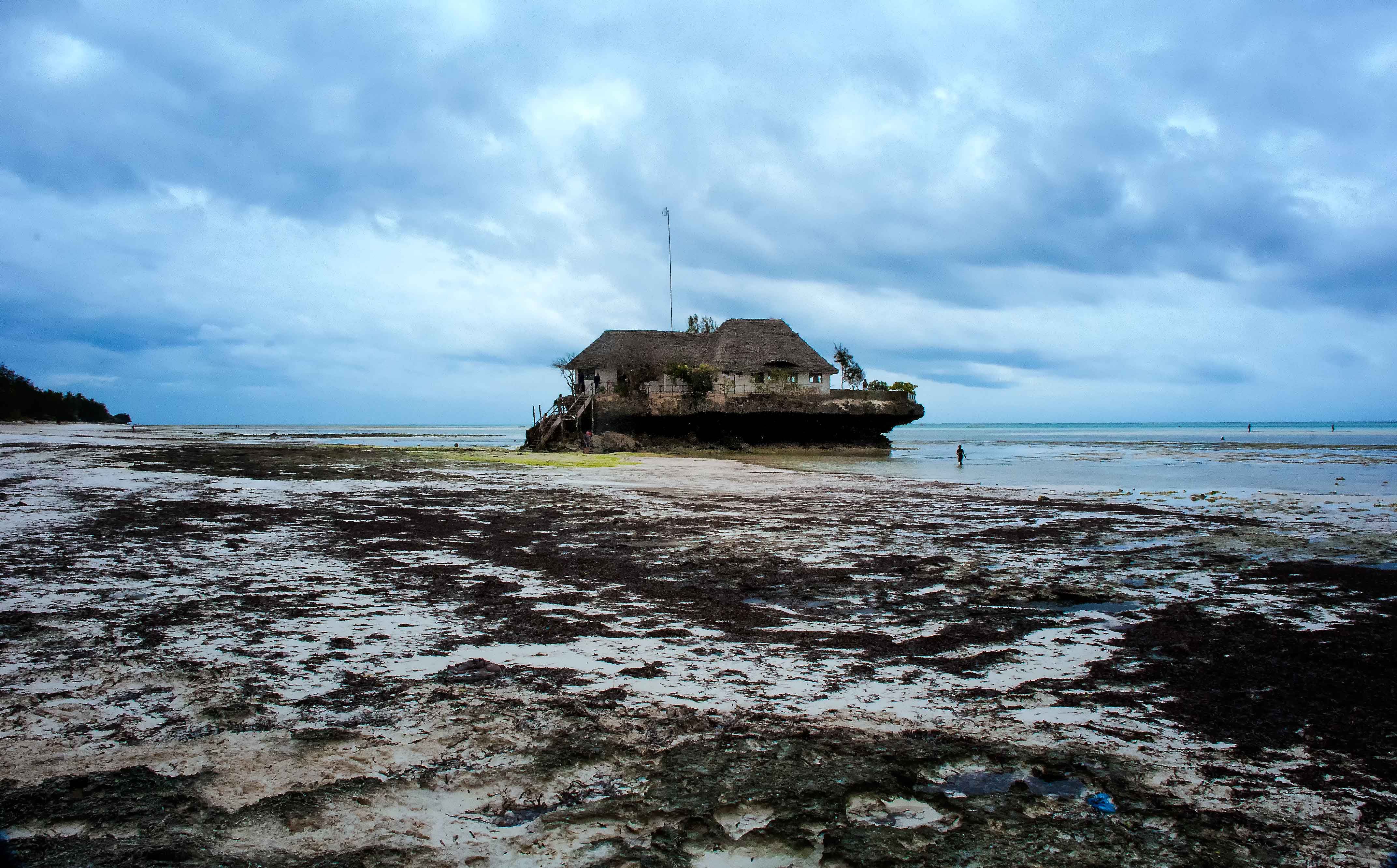 The Rock Restaurant on its coral outcrop off the Michamvi peninsula at low tide, the wide reef flat exposed across the foreground under a dramatic overcast sky, on Zanzibar's southeast coast.